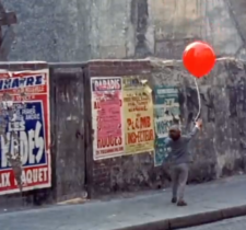 A child holds a bright red balloon against a grey wall plastered with colorful advertising posters.